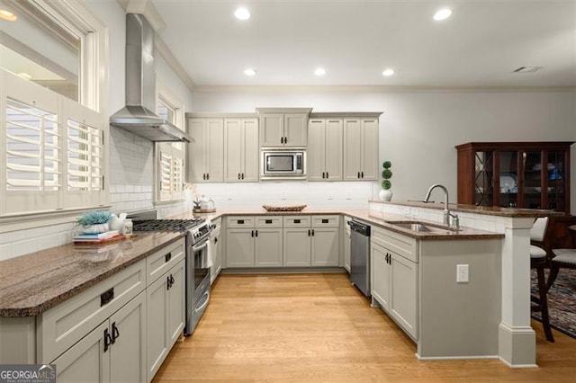 kitchen featuring a peninsula, a breakfast bar area, wall chimney range hood, a sink, and stainless steel appliances