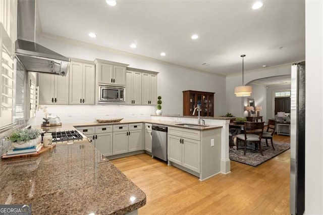 kitchen with a peninsula, wall chimney exhaust hood, a sink, light wood-style flooring, and stainless steel appliances