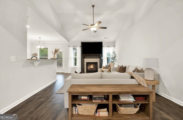living area with lofted ceiling, a lit fireplace, baseboards, dark wood-type flooring, and ceiling fan with notable chandelier