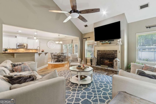 living area featuring visible vents, baseboards, wood finished floors, a stone fireplace, and ceiling fan with notable chandelier