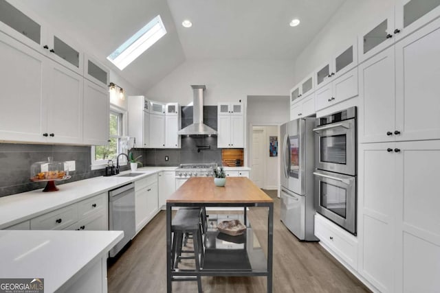kitchen with a sink, wall chimney range hood, a skylight, white cabinets, and appliances with stainless steel finishes