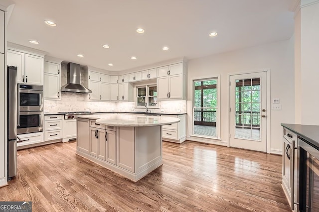 kitchen with appliances with stainless steel finishes, light wood-style flooring, wall chimney exhaust hood, tasteful backsplash, and wine cooler