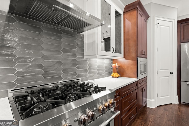 kitchen with wall chimney range hood, backsplash, dark wood-style flooring, appliances with stainless steel finishes, and light countertops