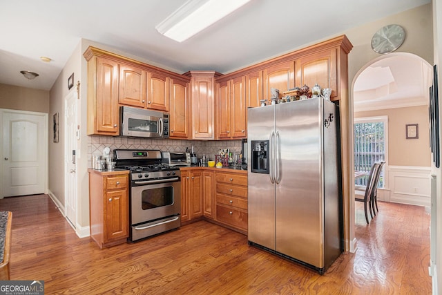 kitchen with tasteful backsplash, light wood-style flooring, appliances with stainless steel finishes, a wainscoted wall, and arched walkways