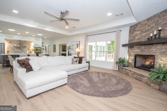 living room featuring ornamental molding, ceiling fan, a stone fireplace, light wood-type flooring, and a raised ceiling