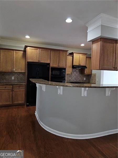 kitchen with decorative backsplash, a breakfast bar area, dark stone counters, brown cabinets, and black appliances