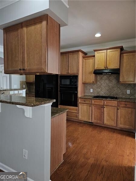 kitchen featuring ornamental molding, wood finished floors, dark stone counters, backsplash, and black appliances
