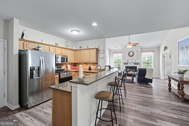 kitchen with dark stone countertops, a breakfast bar area, stainless steel appliances, a warm lit fireplace, and wood finished floors