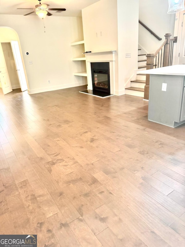 unfurnished living room featuring ceiling fan, arched walkways, a glass covered fireplace, light wood-style flooring, and stairway