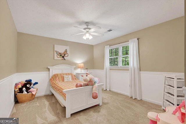 carpeted bedroom with a ceiling fan, a wainscoted wall, and visible vents