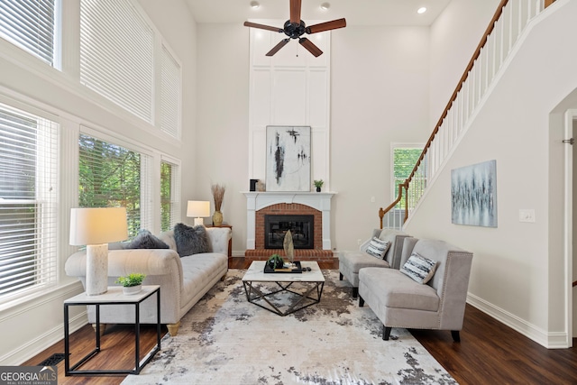 living area with a ceiling fan, a fireplace, stairway, a high ceiling, and dark wood-type flooring
