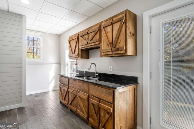 kitchen with brown cabinetry, a paneled ceiling, a sink, baseboards, and dark stone counters