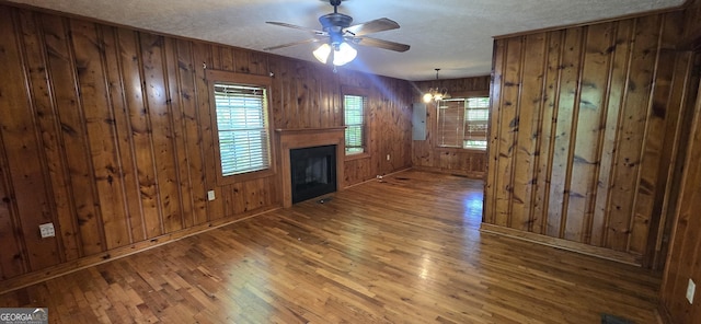 unfurnished living room with electric panel, a fireplace, wood walls, hardwood / wood-style flooring, and ceiling fan with notable chandelier
