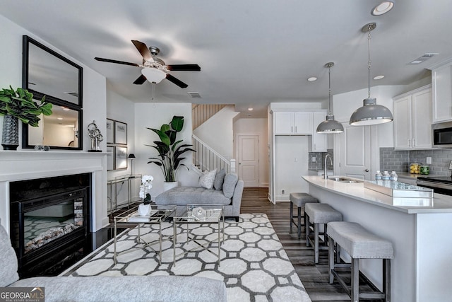living room featuring ceiling fan, visible vents, stairway, dark wood-style floors, and a glass covered fireplace
