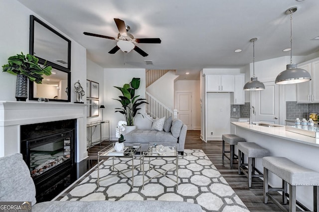 living room featuring ceiling fan, visible vents, a glass covered fireplace, stairs, and dark wood-type flooring