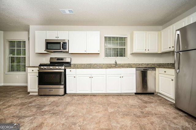 kitchen with a textured ceiling, a sink, dark stone counters, stainless steel appliances, and white cabinetry