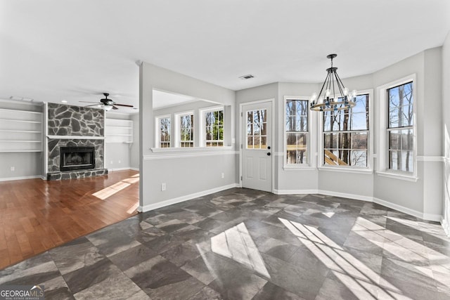 unfurnished dining area with a stone fireplace, baseboards, built in shelves, and ceiling fan with notable chandelier