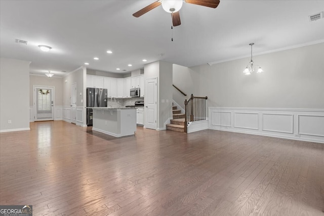 unfurnished living room featuring crown molding, ceiling fan with notable chandelier, a decorative wall, and stairway