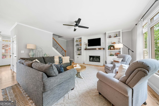 living room with ornamental molding, stairs, a brick fireplace, built in shelves, and a ceiling fan