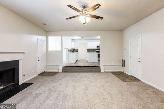 unfurnished living room with a ceiling fan, visible vents, a fireplace with flush hearth, carpet floors, and baseboards