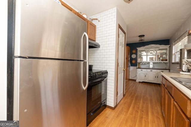 kitchen with wall chimney range hood, black / electric stove, brown cabinets, freestanding refrigerator, and light wood-style flooring