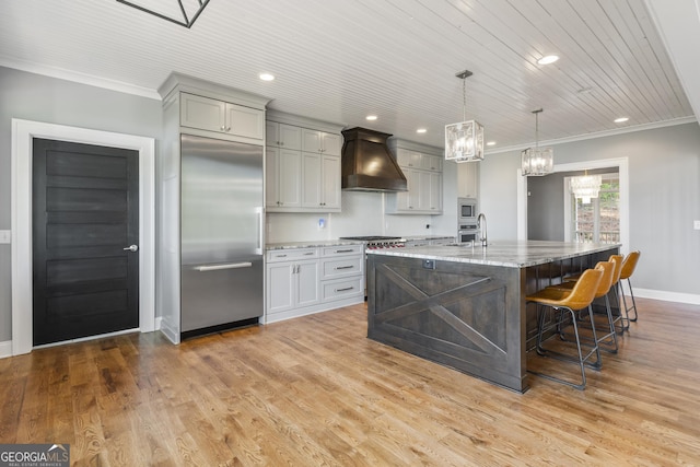 kitchen with light stone counters, built in appliances, a chandelier, ornamental molding, and custom exhaust hood