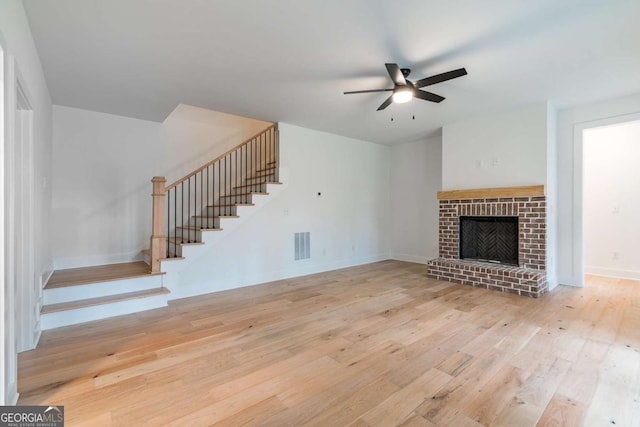 unfurnished living room featuring visible vents, a brick fireplace, wood finished floors, stairway, and a ceiling fan