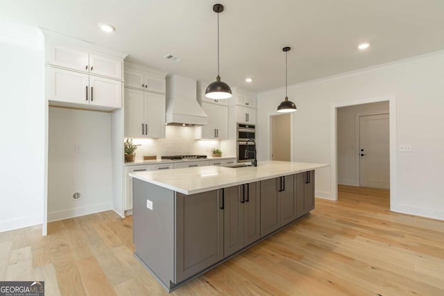 kitchen featuring decorative backsplash, a center island with sink, light countertops, white cabinets, and custom range hood