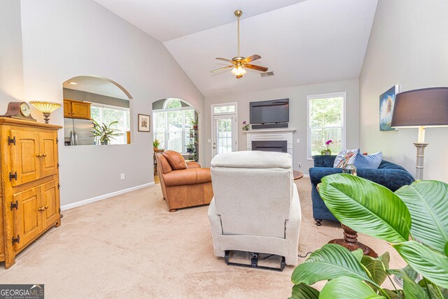 living area featuring a ceiling fan, a fireplace, high vaulted ceiling, light carpet, and baseboards