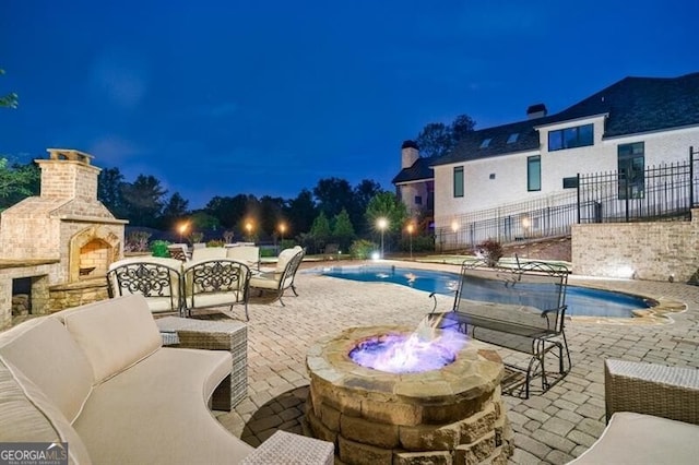 pool at night with fence, an outdoor living space with a fireplace, and a patio area