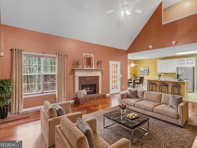living area with light wood-style floors, high vaulted ceiling, a ceiling fan, and a brick fireplace