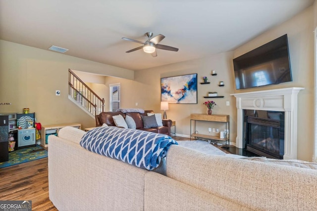 living area with ceiling fan, visible vents, wood finished floors, a glass covered fireplace, and stairs