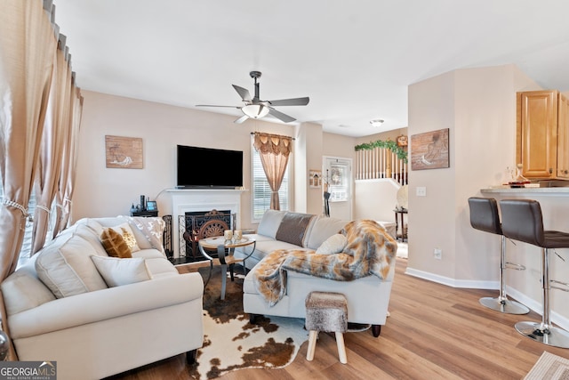 living room with baseboards, ceiling fan, a lit fireplace, and light wood-style flooring