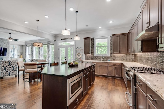 kitchen featuring stainless steel appliances, under cabinet range hood, decorative backsplash, dark wood-type flooring, and open floor plan