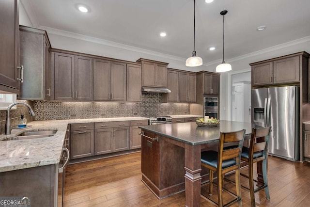 kitchen featuring appliances with stainless steel finishes, a sink, under cabinet range hood, a center island, and light stone countertops
