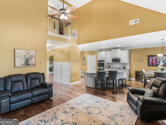 living area with visible vents, light wood-style flooring, a towering ceiling, and ceiling fan with notable chandelier
