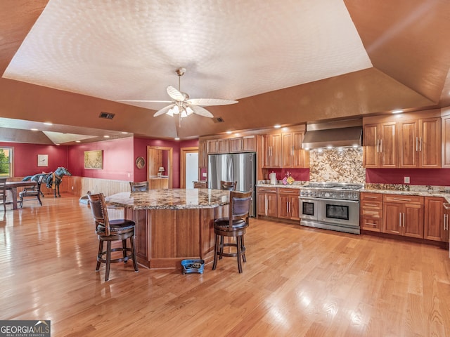 kitchen featuring brown cabinetry, a kitchen island, wall chimney range hood, light stone counters, and appliances with stainless steel finishes