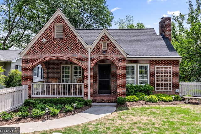 english style home with fence, covered porch, a chimney, and brick siding
