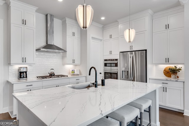 kitchen with a center island with sink, wall chimney range hood, tasteful backsplash, a sink, and appliances with stainless steel finishes
