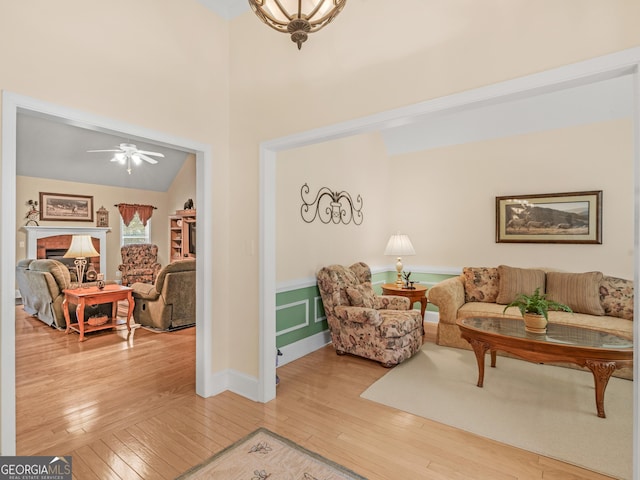 living area featuring light wood-style floors, a ceiling fan, wainscoting, and a brick fireplace