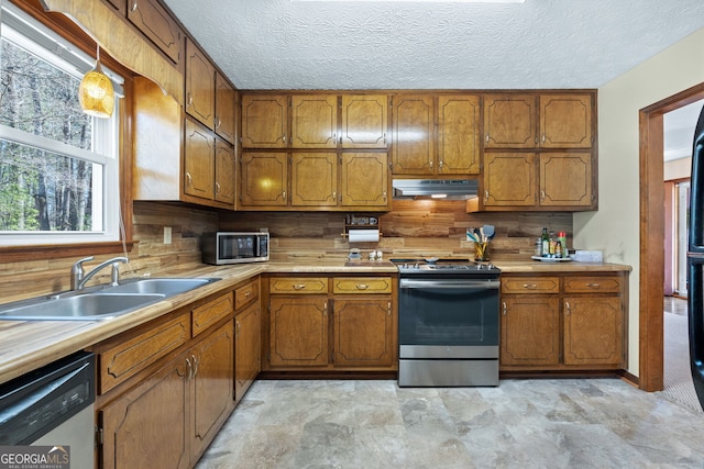 kitchen featuring brown cabinetry, appliances with stainless steel finishes, a sink, light countertops, and under cabinet range hood