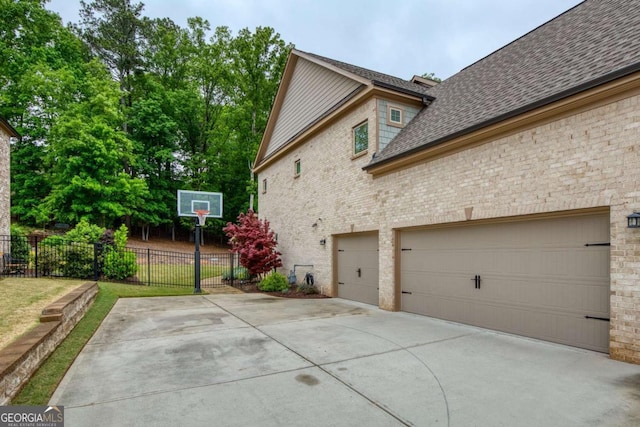 view of home's exterior with driveway, roof with shingles, a garage, brick siding, and fence