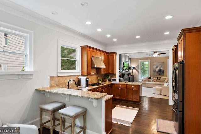 kitchen featuring brown cabinetry, a peninsula, stainless steel gas cooktop, under cabinet range hood, and light stone counters