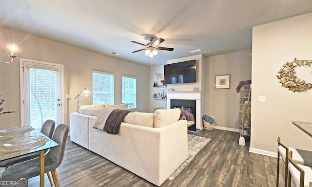 living area featuring a ceiling fan, visible vents, baseboards, dark wood-style floors, and a glass covered fireplace