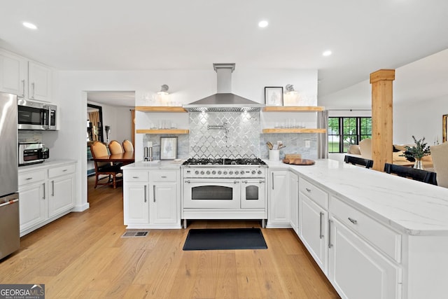 kitchen with open shelves, a peninsula, wall chimney range hood, light wood finished floors, and appliances with stainless steel finishes