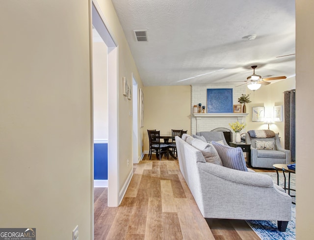 living area with a brick fireplace, a textured ceiling, a ceiling fan, wood finished floors, and visible vents