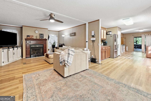living room with ceiling fan, a textured ceiling, light wood-type flooring, a glass covered fireplace, and ornamental molding