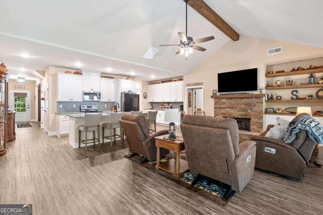 living room with ceiling fan, lofted ceiling with beams, a stone fireplace, wood finished floors, and visible vents