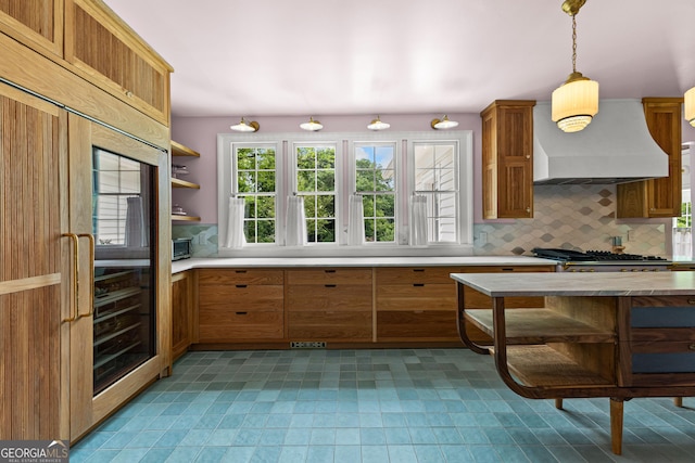kitchen featuring light countertops, brown cabinetry, premium range hood, decorative backsplash, and refrigerator with glass door