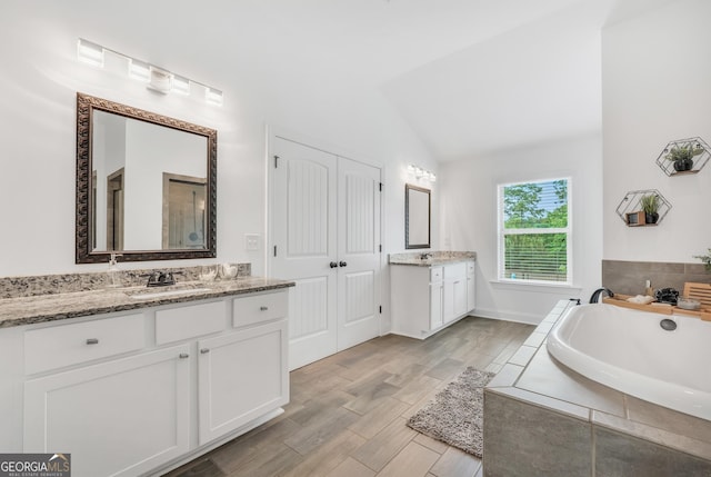 full bathroom featuring a sink, two vanities, wood finished floors, vaulted ceiling, and a garden tub